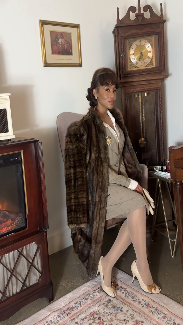 Fur-collar coat over a tweed suit, seated